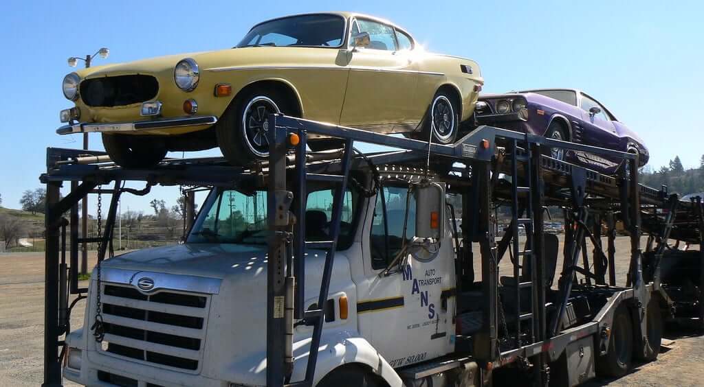 Open trailer with two cars loaded on the top, one is a yellow Volvo P100 2 door coupe and the other is a purple American muscle car. Open car shipping is the most common way to transport cars. Open car shipping or enclosed trailer, National Transport Services provides both auto transport services from coast to coast.