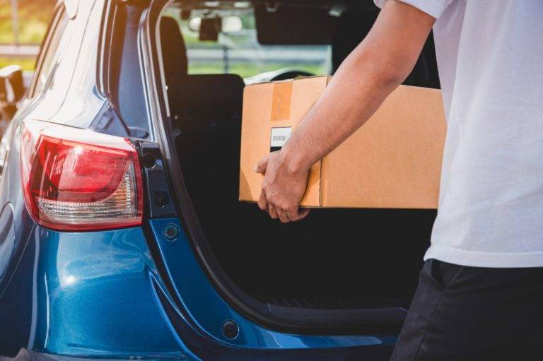 A man puts a cardboard box in his trunk. Ship a car with stuff inside.