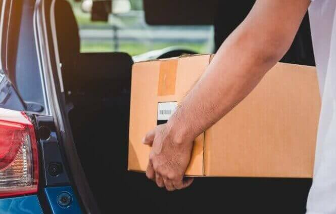 A man puts a cardboard box in his trunk. Ship a car with stuff inside.