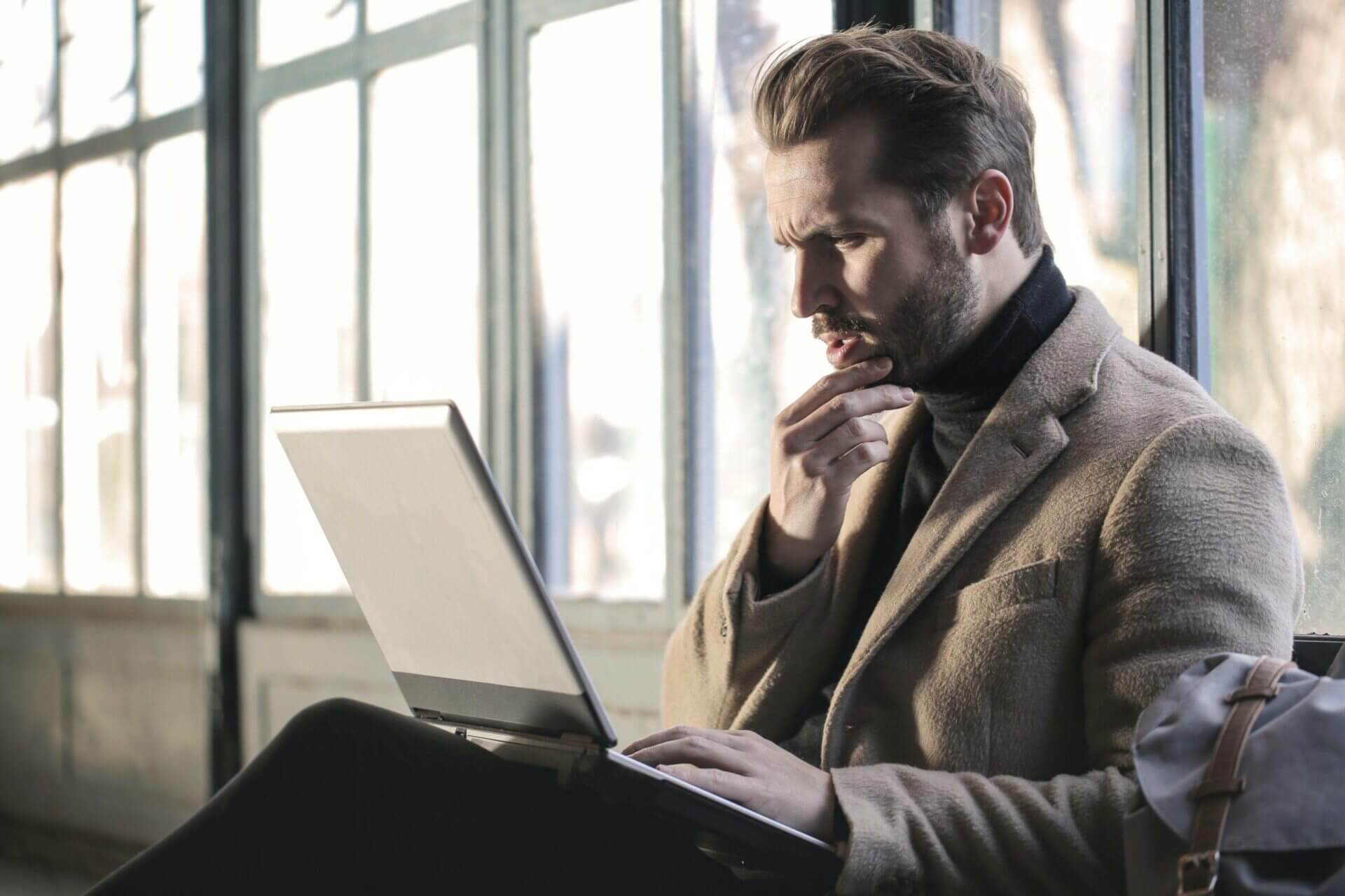 A man sitting wearing an overcoat looking at his laptop. 