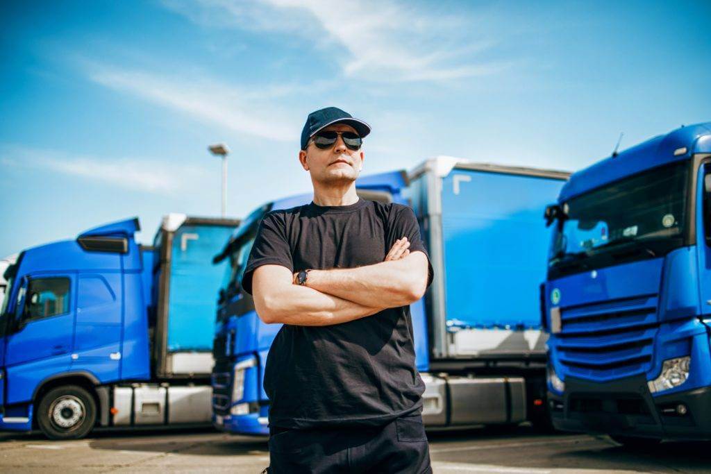 A car carrier driver is standing in front of his truck with his arms crossed. Only the very best car shipping companies vet each and every car carrier.