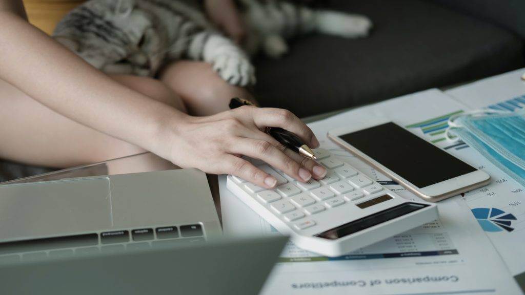 A hand holding a pen while typing on a calculator with a cell phone and laptop on a table. Subprime borrowers are facing difficulties making payments. Car insurance and car payments are becoming harder to manage. New car supply and vehicle prices are coming down. Consider trading for a cheaper car to lower your monthly payments.