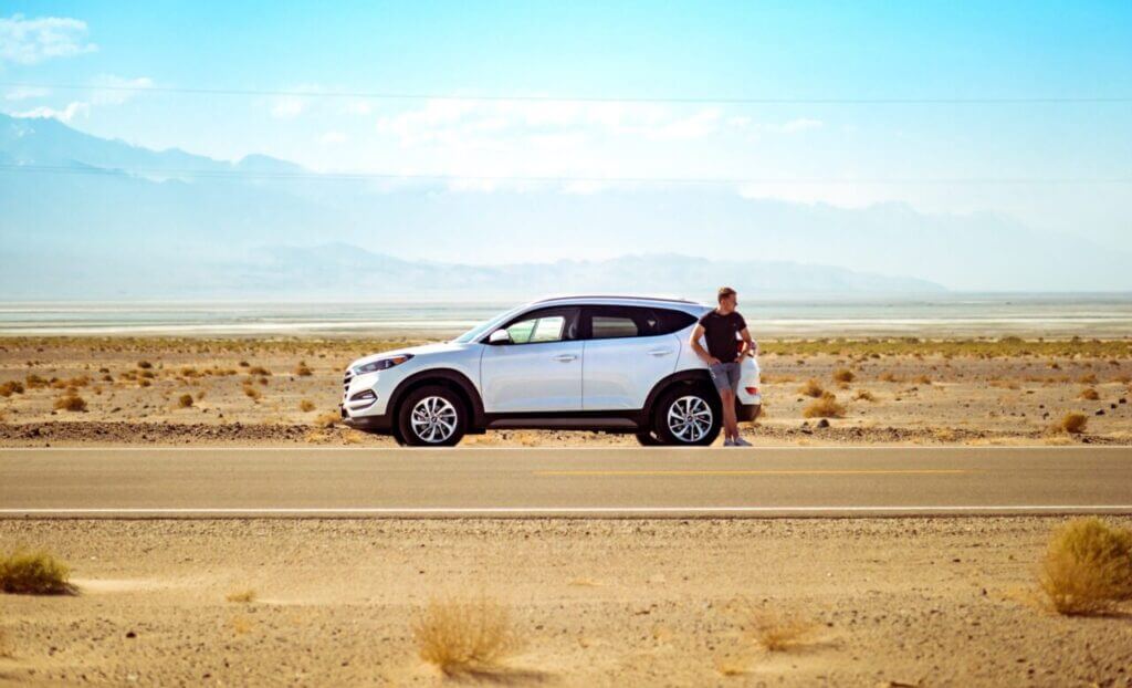 A man is standing outside his vehicle, stranded on a desert road. It might not be a good idea to drive your car cross country.
